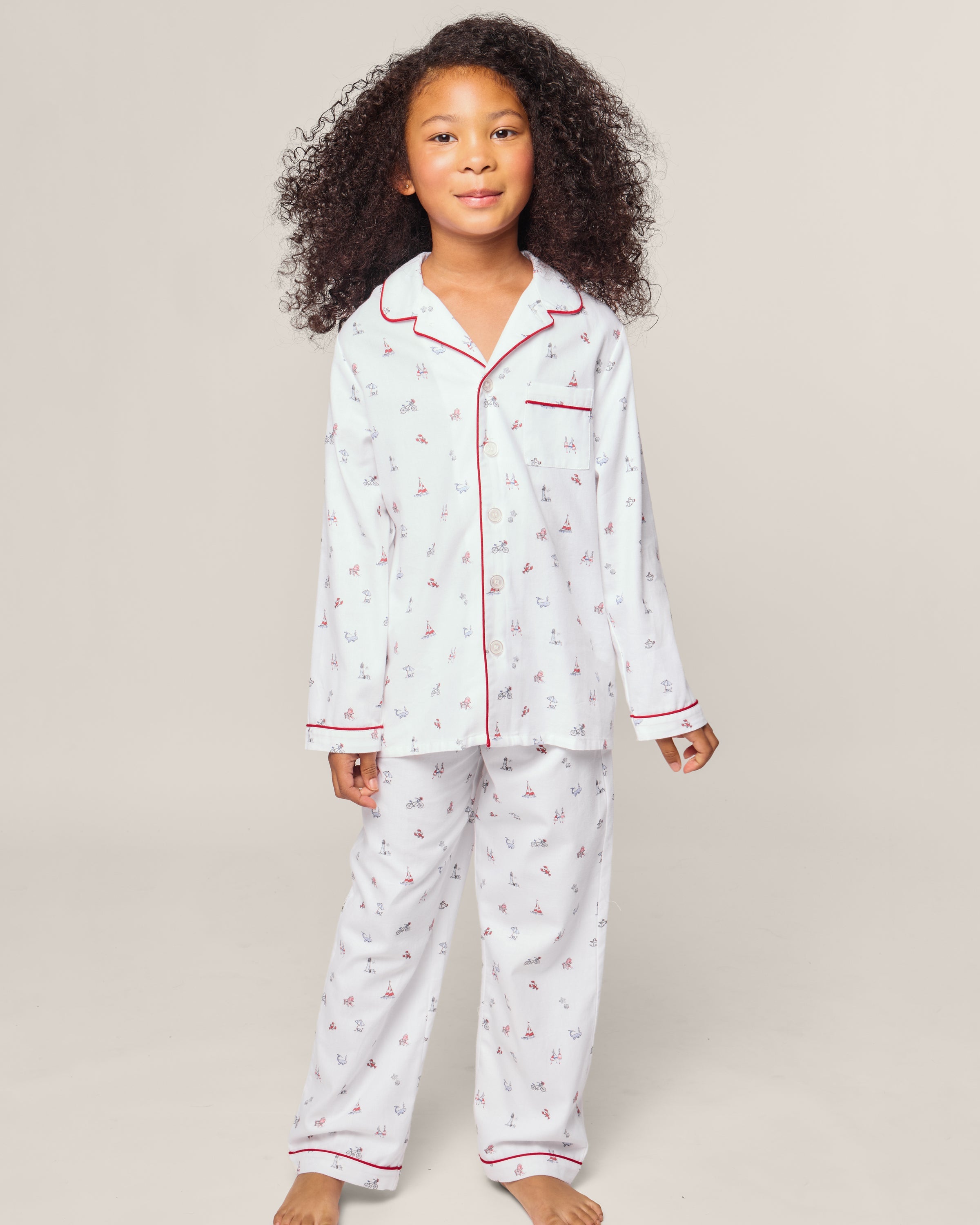 A child with curly hair stands in Petite Plumes Kids Twill Pajama in Coastal Charms—a charming, flame retardant set with a long-sleeve top and pants featuring small designs on a white background with red trim.