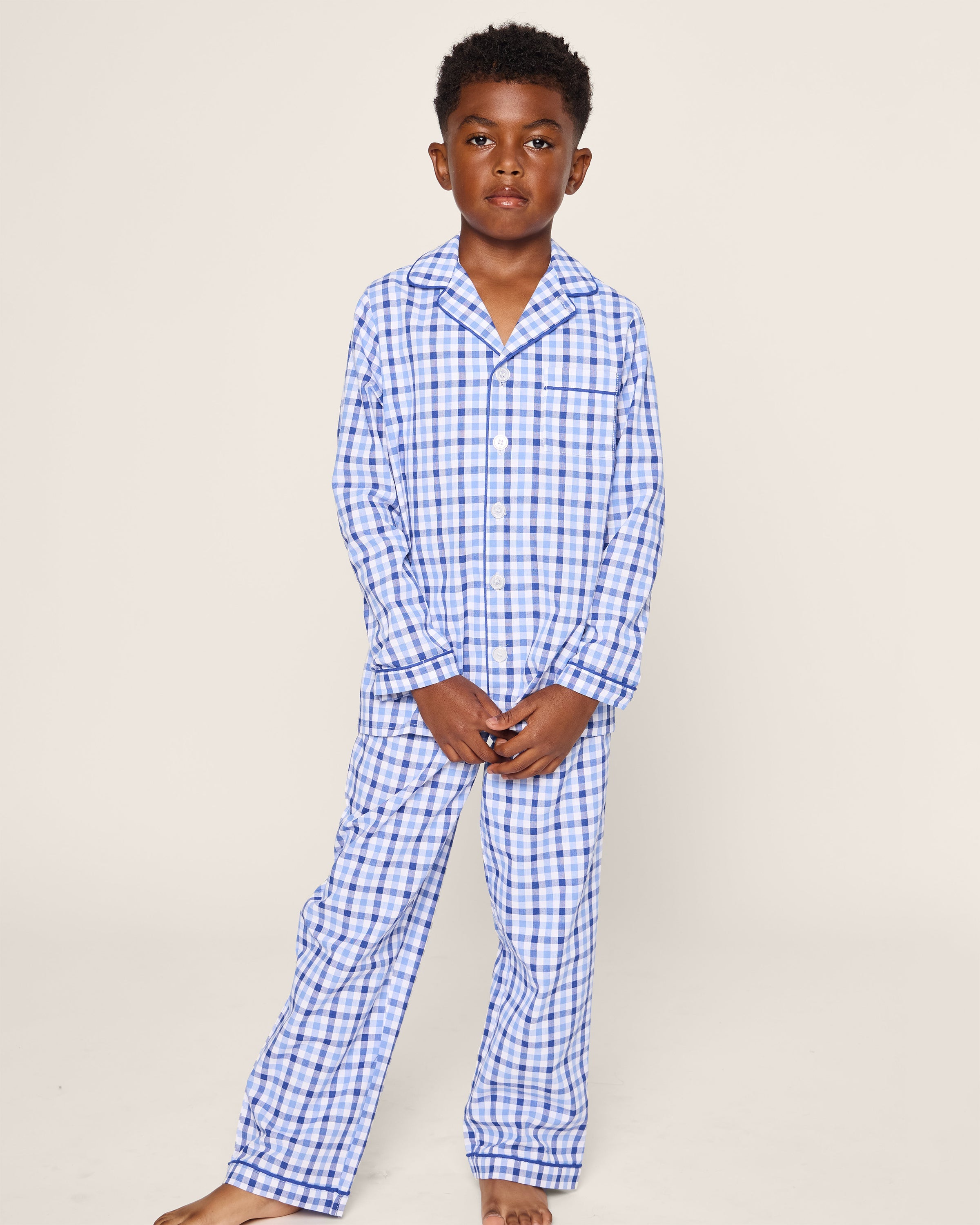 A young boy sports Petite Plumes Kids Twill Pajama Set in Royal Blue Gingham. He is barefoot with short, curly hair, and a plain background highlights his luxury sleepwear’s gingham print.
