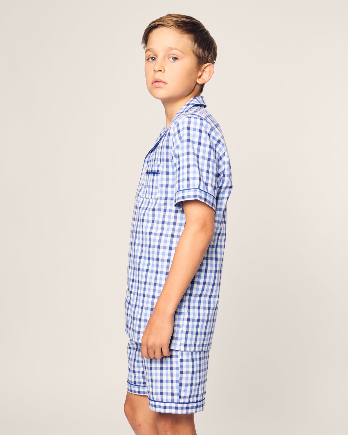 A young boy stands sideways, looking at the camera in a Kids Twill Pajama Short Set by Petite Plume. He wears a Royal Blue Gingham yarn-dyed cotton shirt and matching shorts against a plain light-colored background.