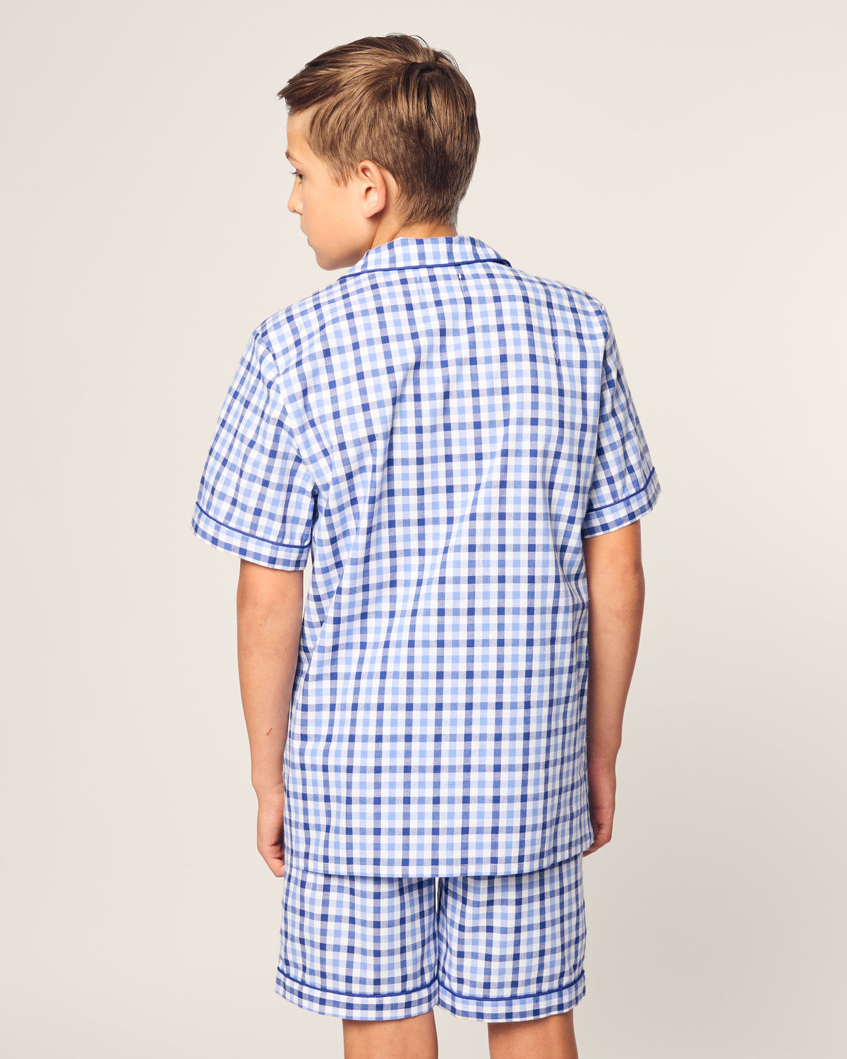 A young boy faces away, dressed in Petite Plumes Kids Twill Pajama Short Set in Royal Blue Gingham, made of yarn-dyed cotton with short sleeves and matching shorts. The background is plain off-white.