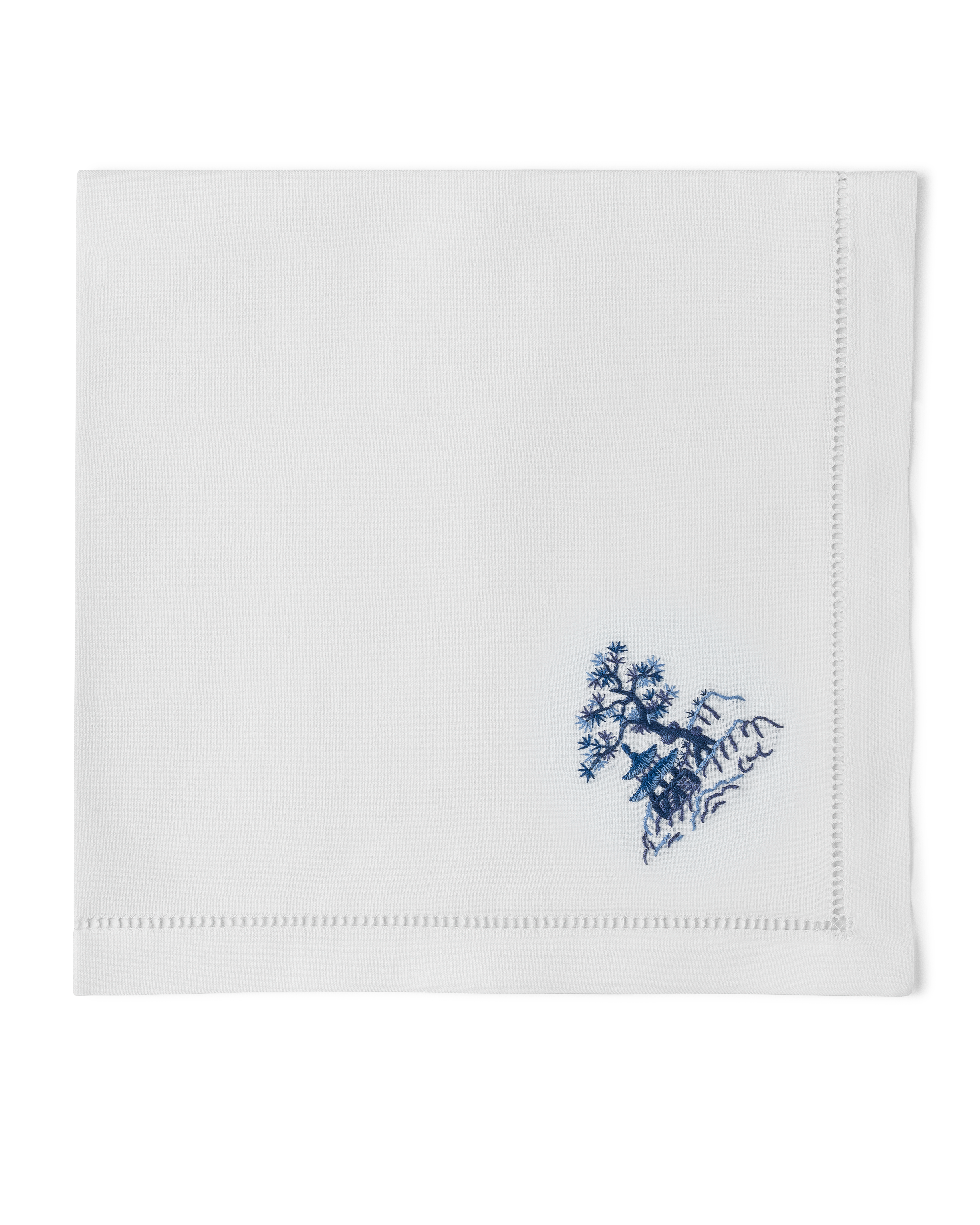 A white napkin with a hemstitch. A house & tree on a hill are embroidered in blue in the bottom righthand corner.