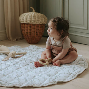 Secret Garden play mat in Ivory. Child is sitting on play mat with stuffed animal and rattle