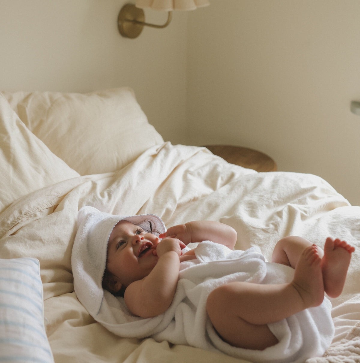 Baby hooded towel in white with blue trim detail wrapped around baby laying in bed with feet playfully up in the air