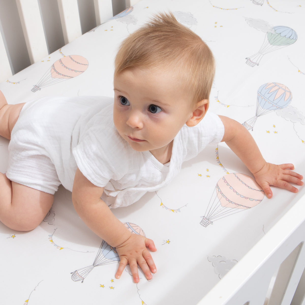 Crib Sheet in "Touch The Sky" print in the color pink featured in a crib with baby doing tummy time 