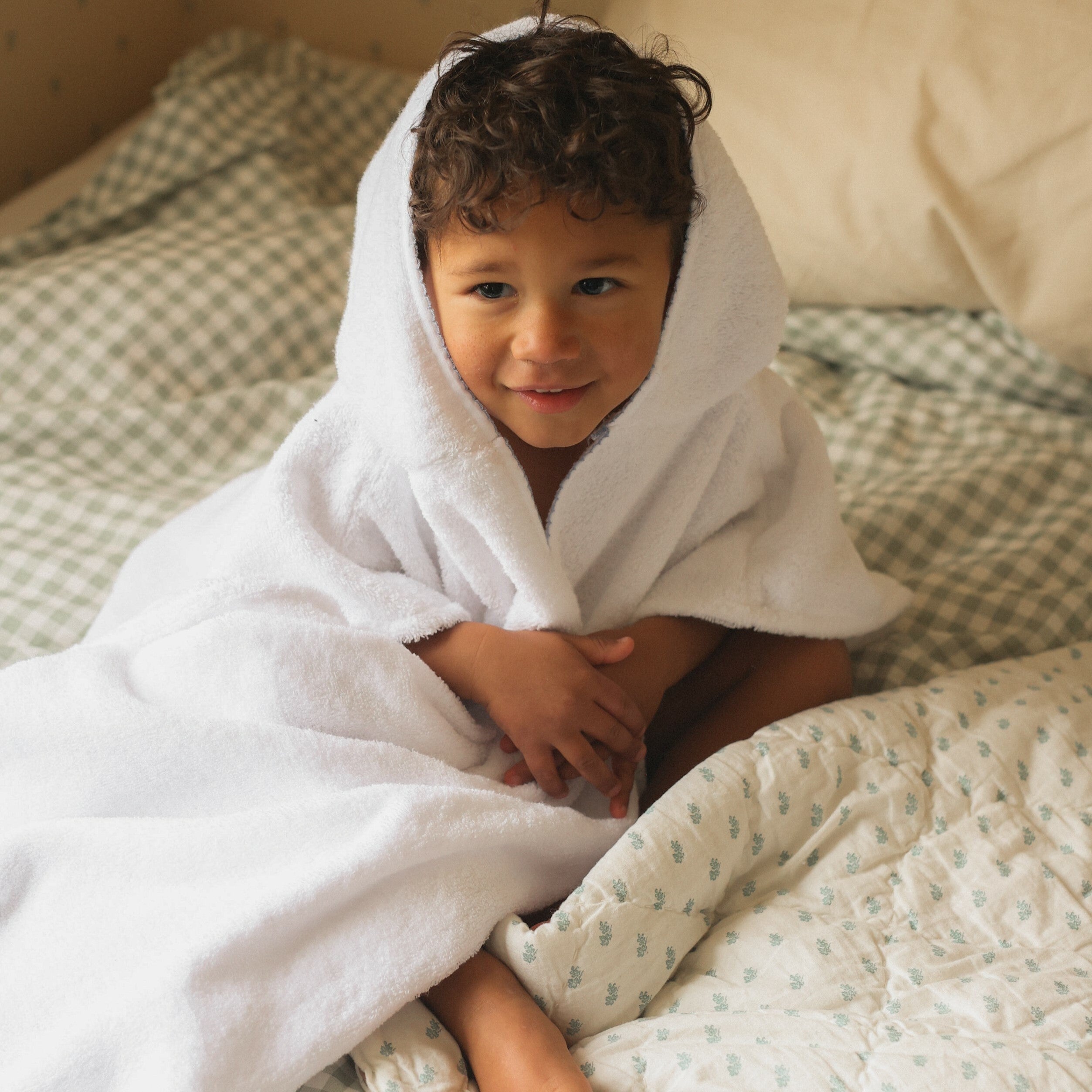 Toddler Poncho in white with blue trim detail. Child is wrapped in Toddler poncho with hood on his head. Sitting in a cozy bed