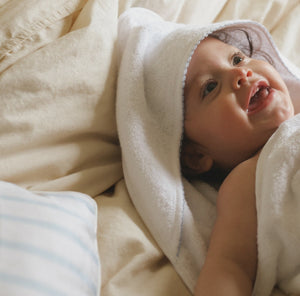 Baby Hooded Towel in white with blue trim detail. Baby is wrapped in hooded towel laying on a bed smiling 